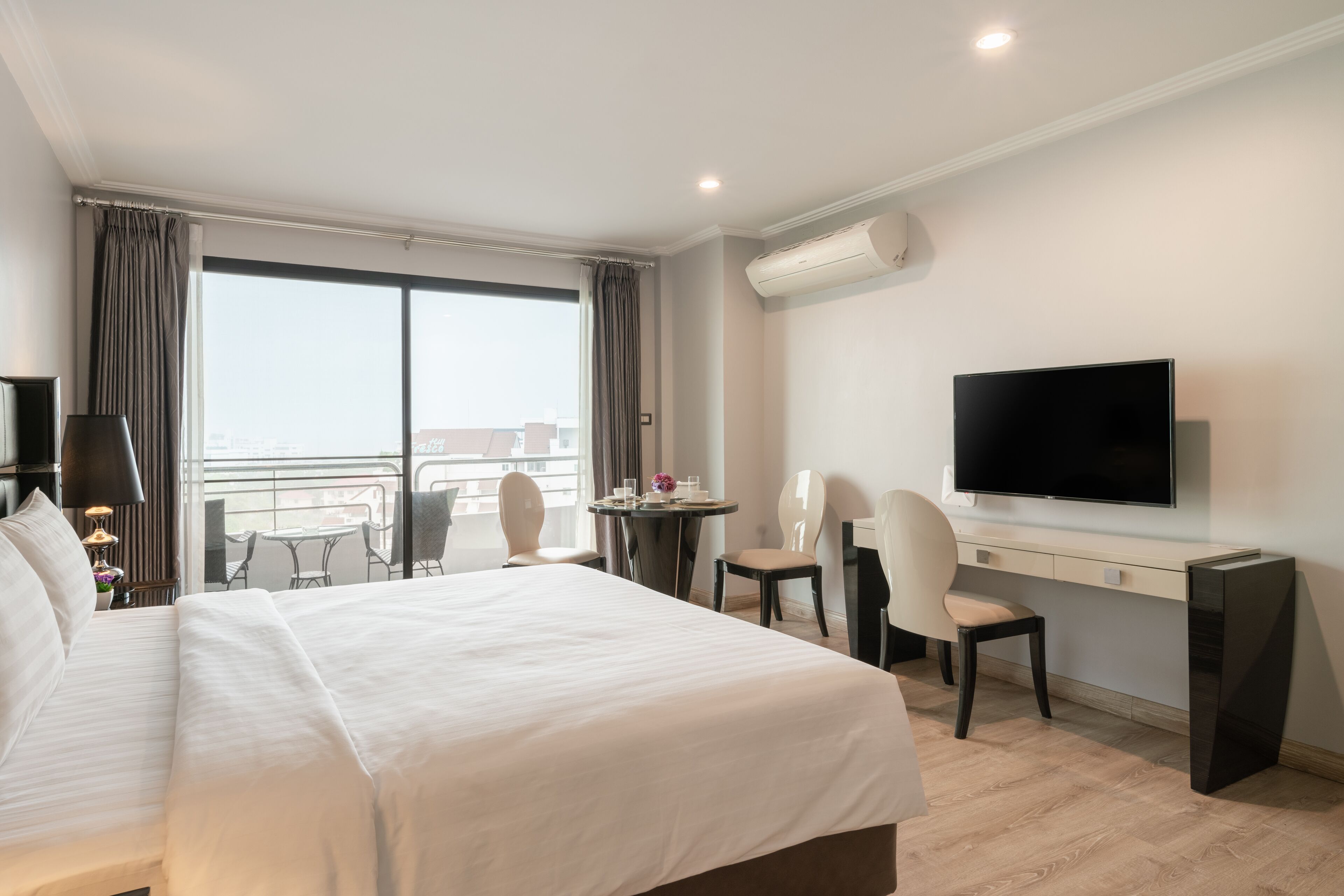Et hotelværelse med en stor seng, et skrivebord med et fjernsyn, en balkon med et bord og stole samt en aircondition.