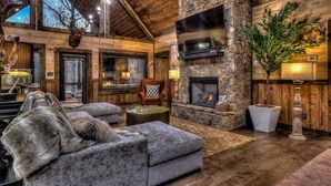 A log cabin interior with a stone fireplace, a mounted television, and a cozy seating area with a fur throw. - Luxe Mountain Retreat - Pet Friendly | Multiple Outdoor Living Areas | Hot Tub | Game Room (Blue Ridge)