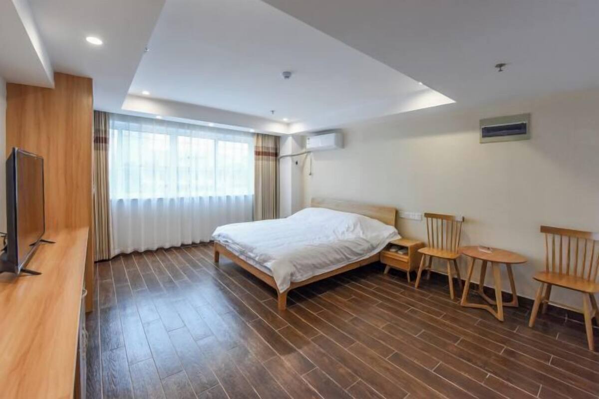 A hotel room with a wooden floor, a bed with white bedding, a TV on the left, and two wooden chairs near the window.