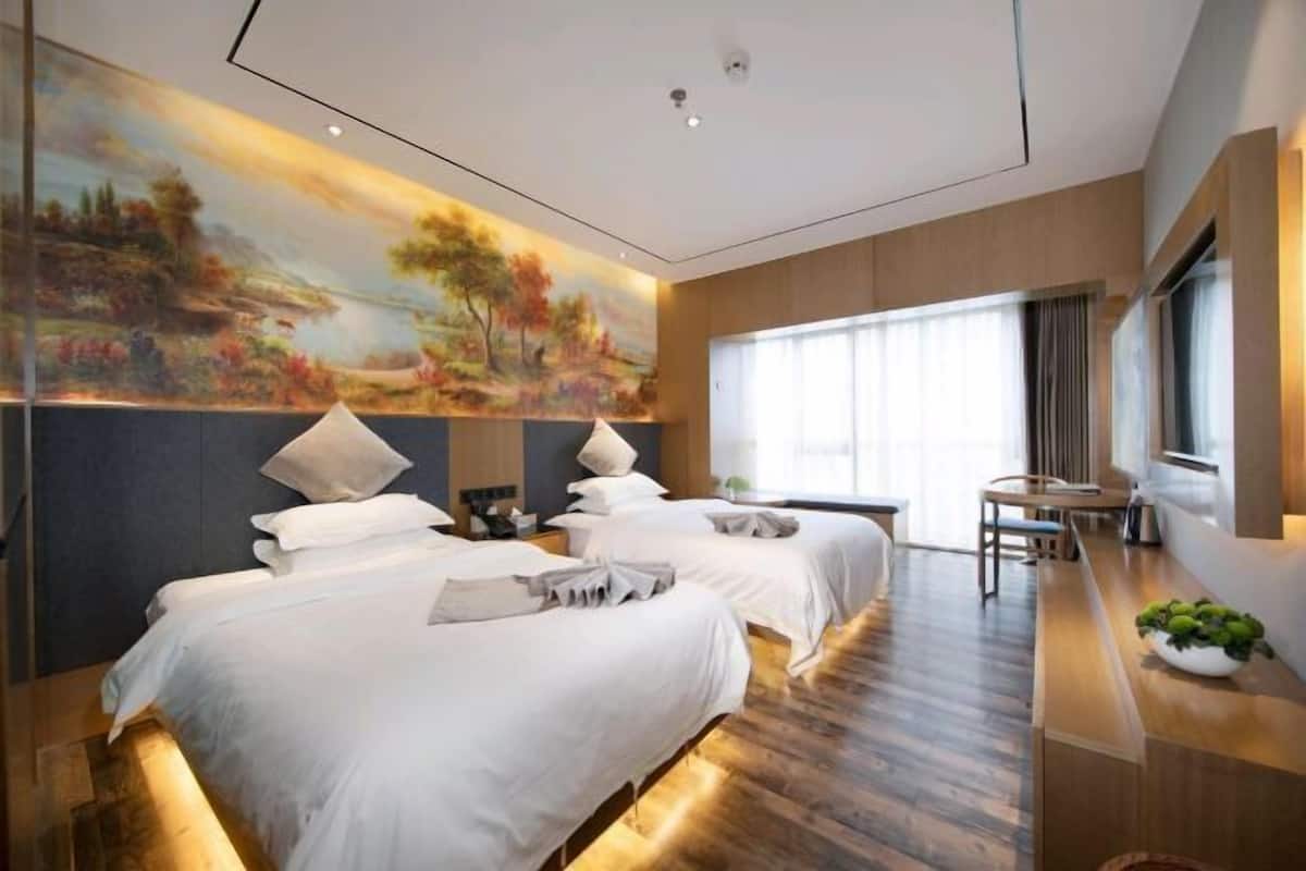 A hotel room with two beds, a painting on the wall, a desk with a chair, and a large window.