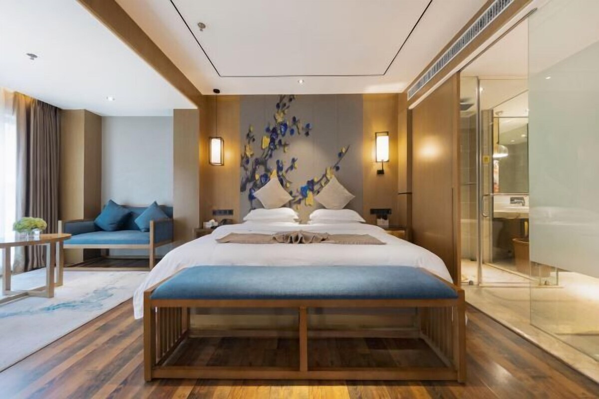 Quarto de hotel moderno com uma cama grande, um sofá, uma mesa pequena e um objeto decorativo na parede.