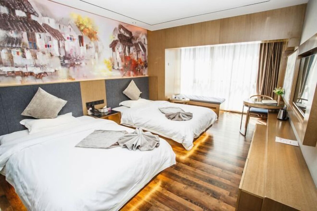 Quarto de hotel com duas camas, piso de madeira, uma janela grande com cortinas e um quadro na parede.