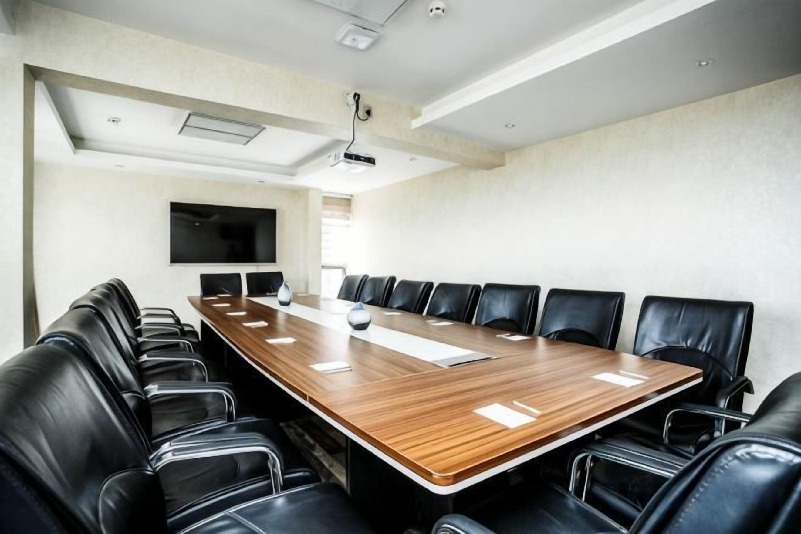 A conference room with a long wooden table, black leather chairs, a flat-screen TV, and a projector mounted on the ceiling.