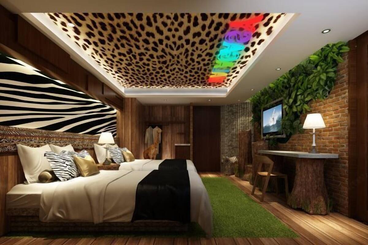 A bedroom with a zebra-patterned headboard, a bed with a black throw, a wooden ceiling with a leopard print design, a TV mounted on a brick wall, and a small desk with a lamp.
