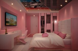 Un dormitorio moderno con una decoración en tonos rosados, una cama con ropa de cama blanca, un escritorio con una silla y un cuadro en la pared.