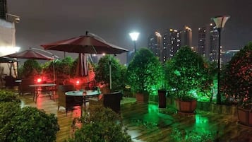 Un patio al aire libre con mesas y sombrillas, rodeado de plantas en macetas e iluminado con luz ambiental.