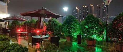 An outdoor patio area with tables and umbrellas, surrounded by potted plants and illuminated by ambient lighting.