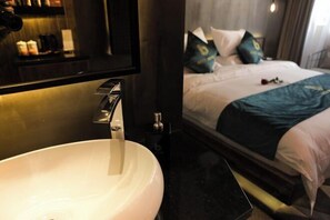 Habitación de hotel con un lavamanos grande y blanco, una cama con ropa de cama verde azulado y blanca, y un espejo que refleja la habitación.