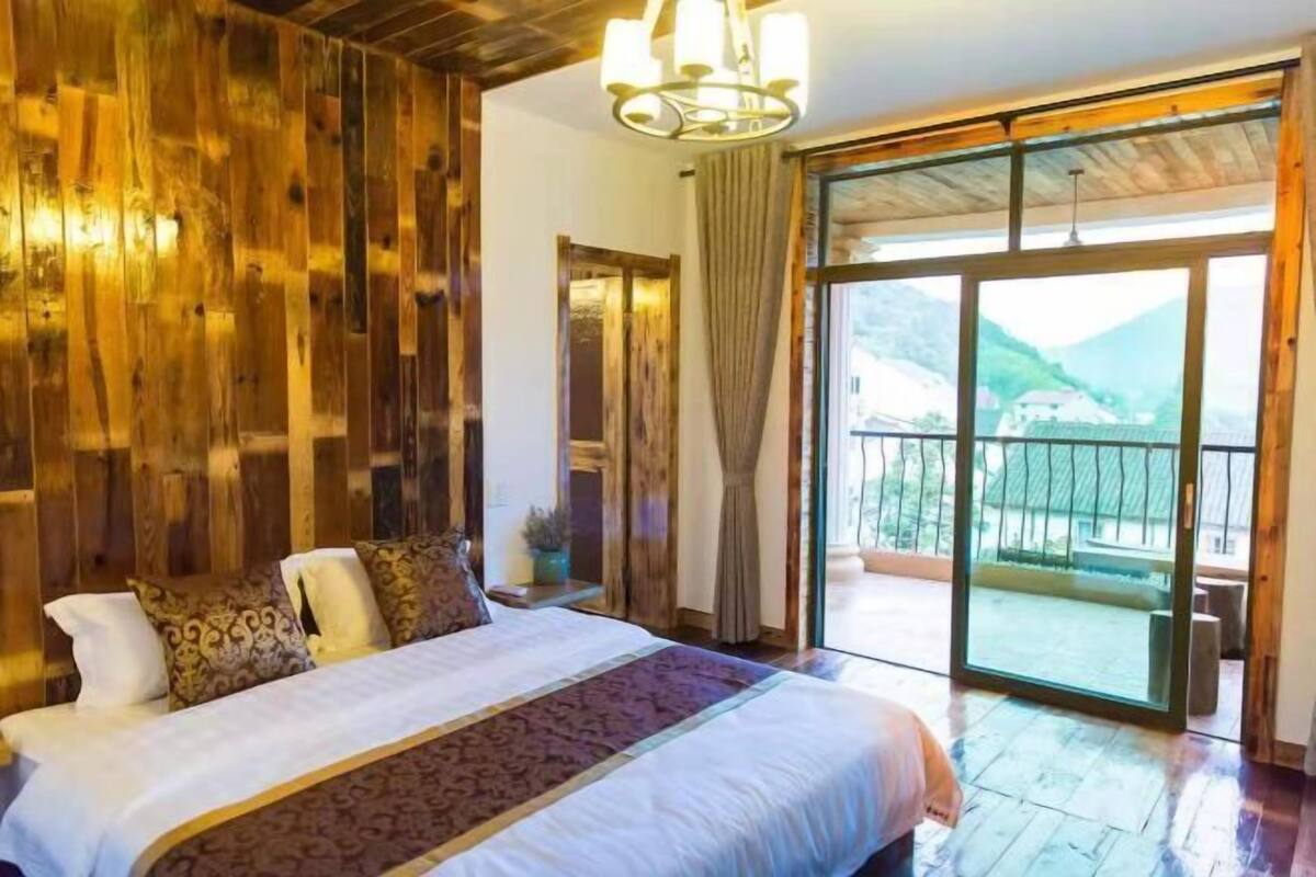 A bedroom with a large bed, wooden walls, a balcony with a view of the mountains, and a sliding glass door.