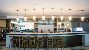 Una zona de bar moderna con una barra central, varios taburetes, iluminación colgante y una pantalla digital.