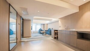 Una habitación de hotel moderna con kitchenette, un área de estar con sofá y sillones, y un gran ventanal que proporciona luz natural.