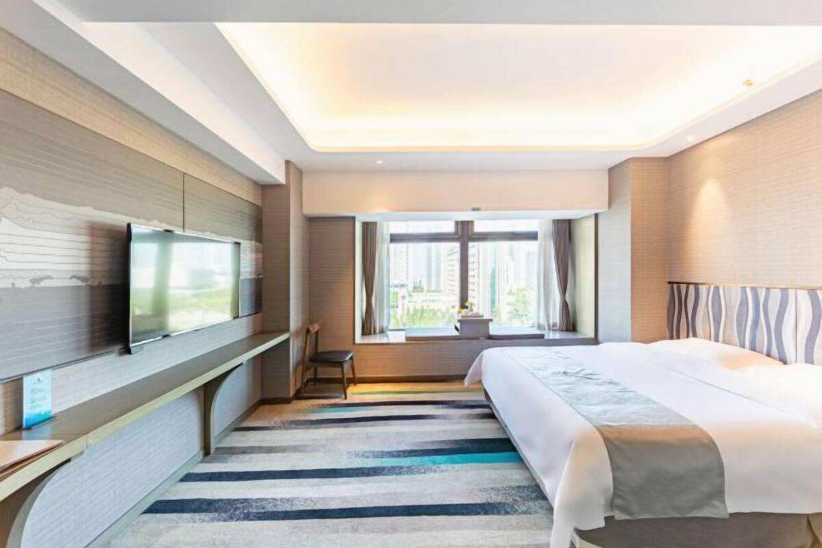 Habitación de hotel con cama, televisión, ventana con cortinas y vistas a edificios.
