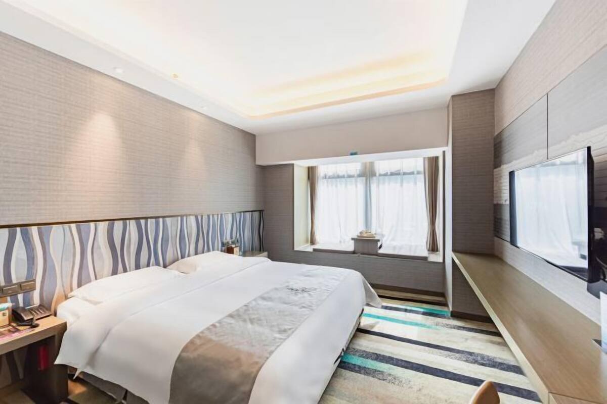 Habitación de hotel con cama, mesita de noche, lámpara y un amplio ventanal con cortinas.