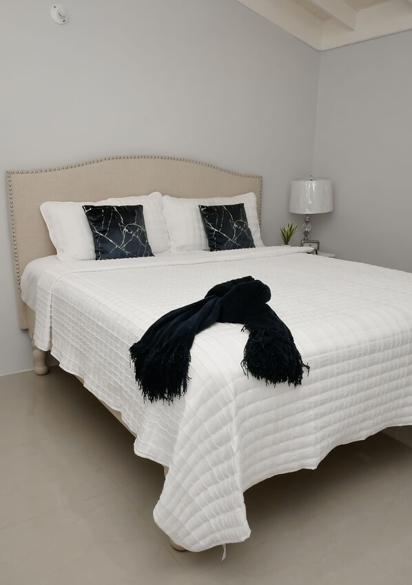 A neatly made bed with a white quilt and a black throw blanket, a beige upholstered headboard, and a bedside lamp. - Vacation Home 5 minutes from the beach (parròquia de Trelawny)