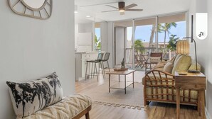 A modern living room with a sofa, a coffee table, and a wicker chair. There is a dining area with chairs and a table. The room has large windows and a view of the outdoors. -  NO PARKING & RESORT FEE IN WEST MAUI WITH AMAZING OCEAN VIEW CONDO (Lahaina)