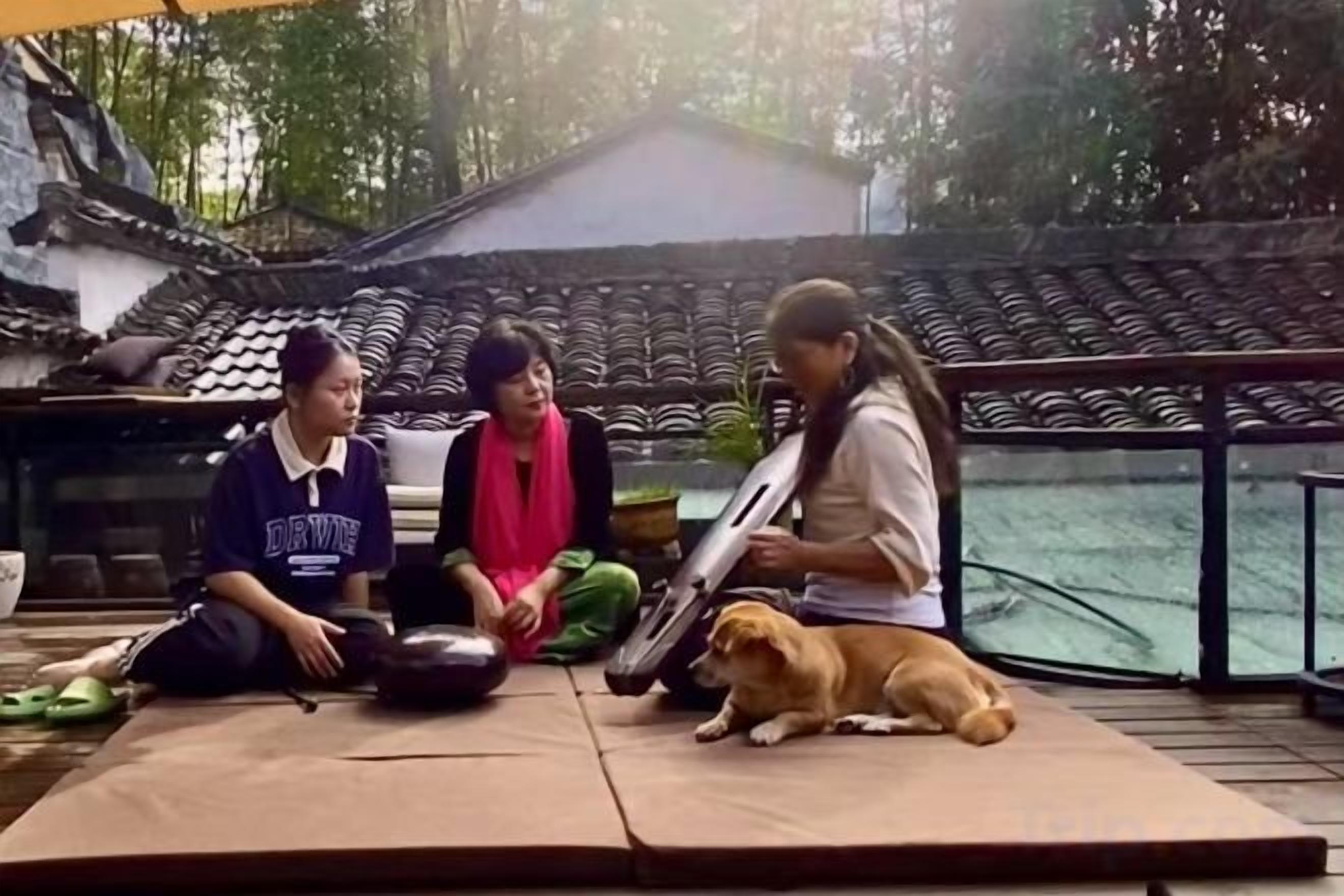 Three individuals sitting on mats outdoors with a dog, near a traditional building with tiled roofs.