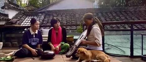 Three individuals sitting on mats outdoors with a dog, near a traditional building with tiled roofs.