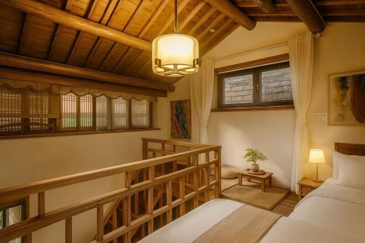 Una habitación con techo de madera, una cama, una mesita de noche, una lámpara, un ventanal con cortinas y una pequeña mesa con una planta.
