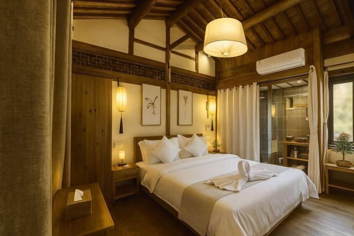 Une chambre d’hôtel avec un plafond en bois, un lit recouvert de draps blancs, des lampes fixées au mur et une fenêtre avec des rideaux.
