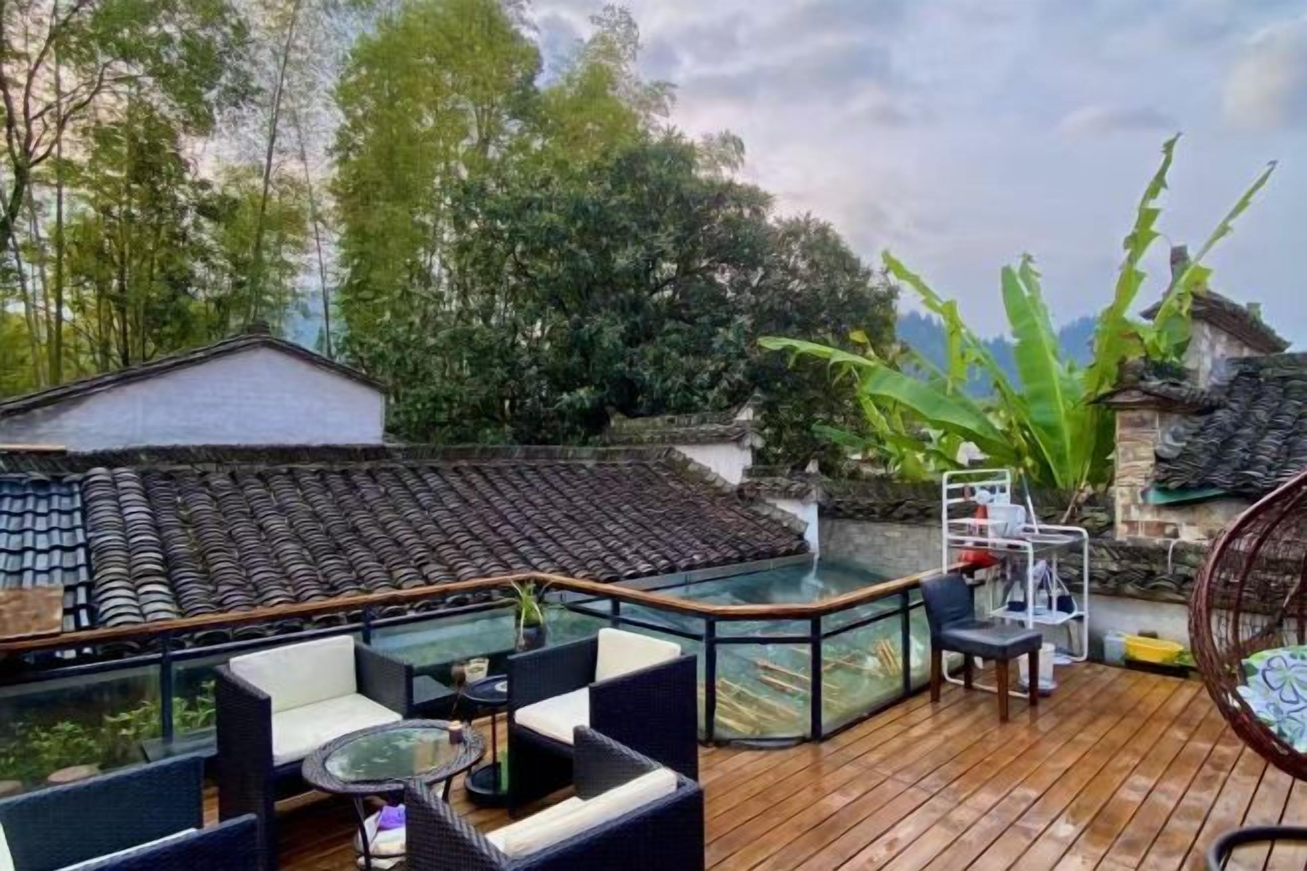 Una terraza de madera con mobiliario exterior, barandilla de cristal y vista a un edificio tradicional y árboles.