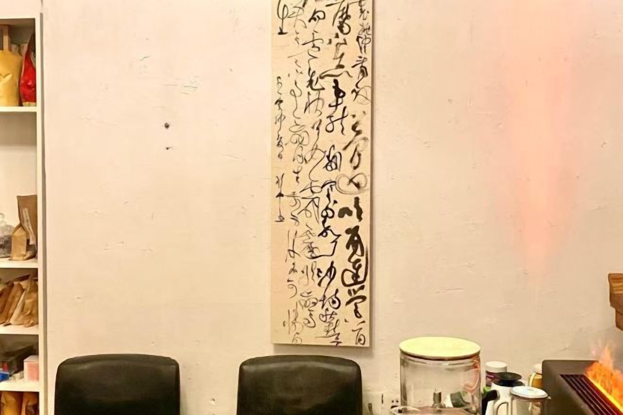 A room with a long wall-mounted artwork, two black chairs, and a small table with a jar and a container.
