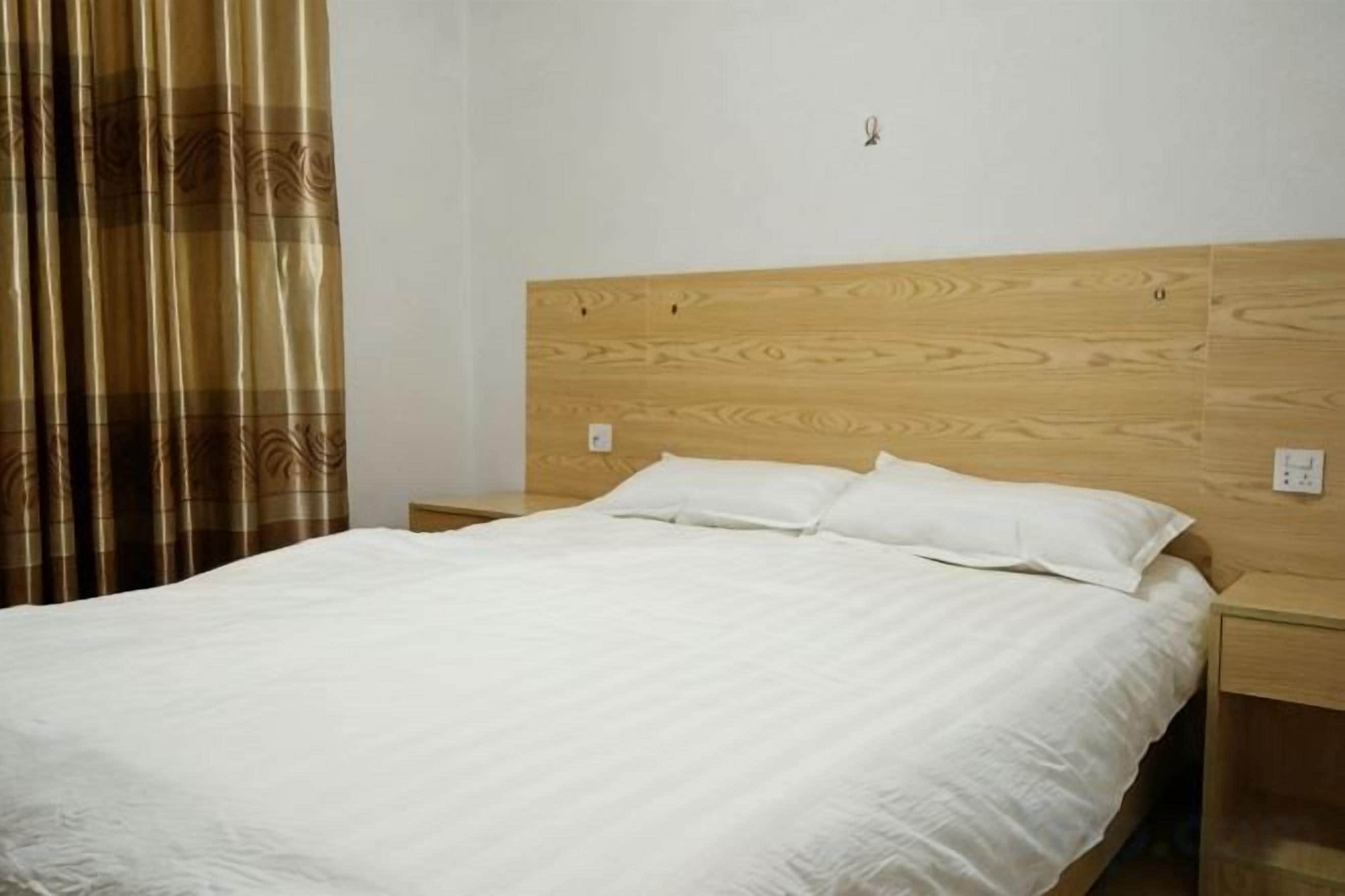 A neatly made bed with a wooden headboard and white bedding in a simple room with a curtain and a wall socket.