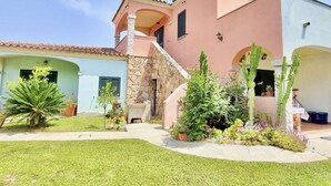 Una casa de dos pisos con una escalera de piedra, un pequeño jardín con diversas plantas y un cielo azul despejado.