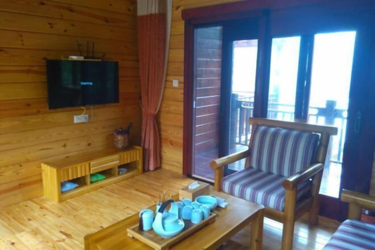 Interior de una cabaña de madera con un televisor montado en la pared, un sofá a rayas, una mesa con vajilla de té y un ventanal corredero que da a un espacio exterior.