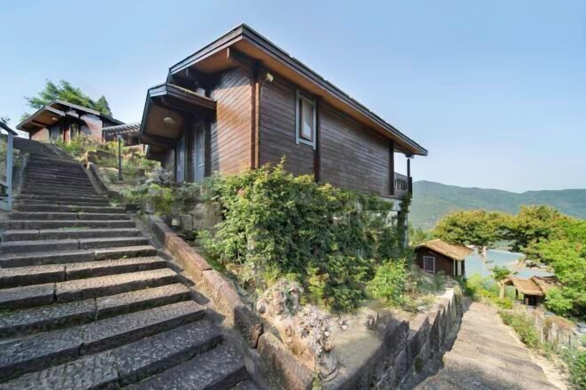 Una casa moderna de madera con una escalera que conduce hasta ella, rodeada de vegetación y vistas a las montañas.