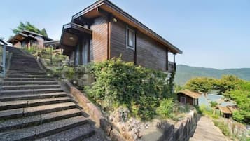 Uma casa moderna de madeira com uma escadaria de pedra íngreme que a leva ao topo, rodeada por vegetação e colinas.