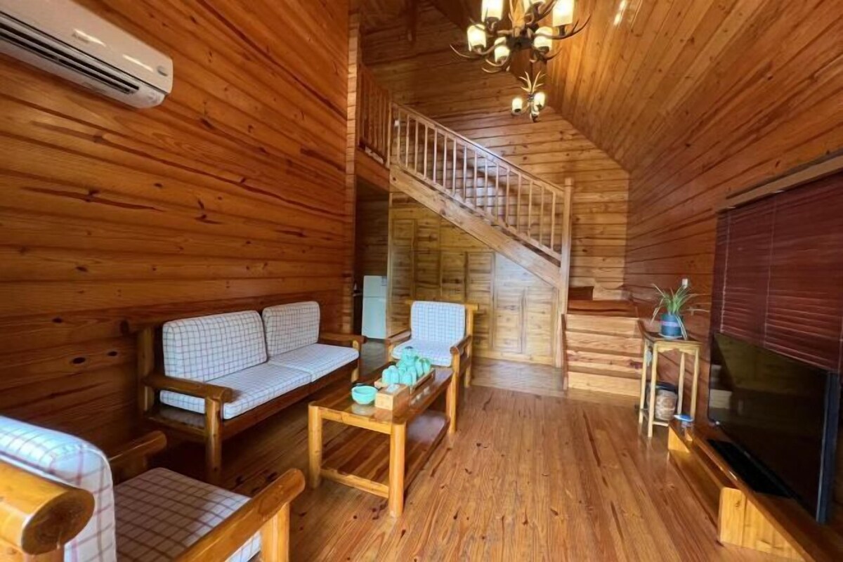 Interior de una cabaña de madera con sofá, mesa, televisión y escalera.