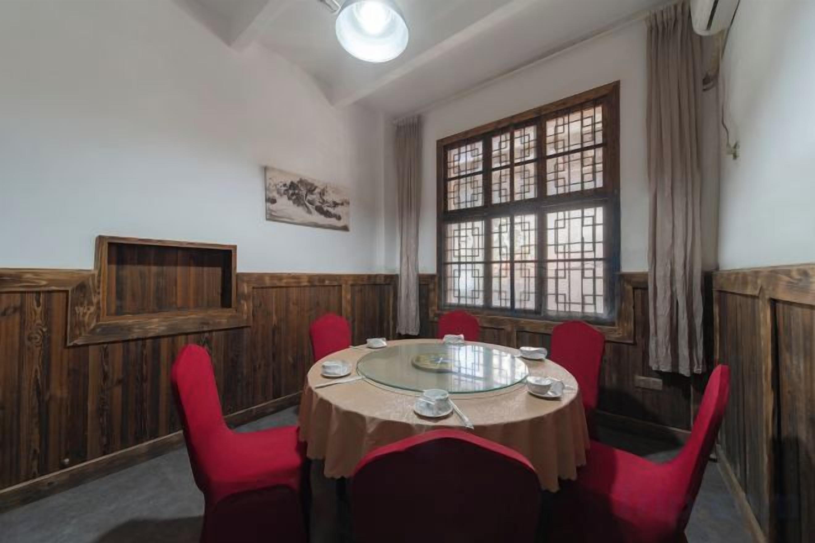 A round table with a glass top surrounded by red chairs, set in a room with wooden paneling and a large window with a grid pattern.