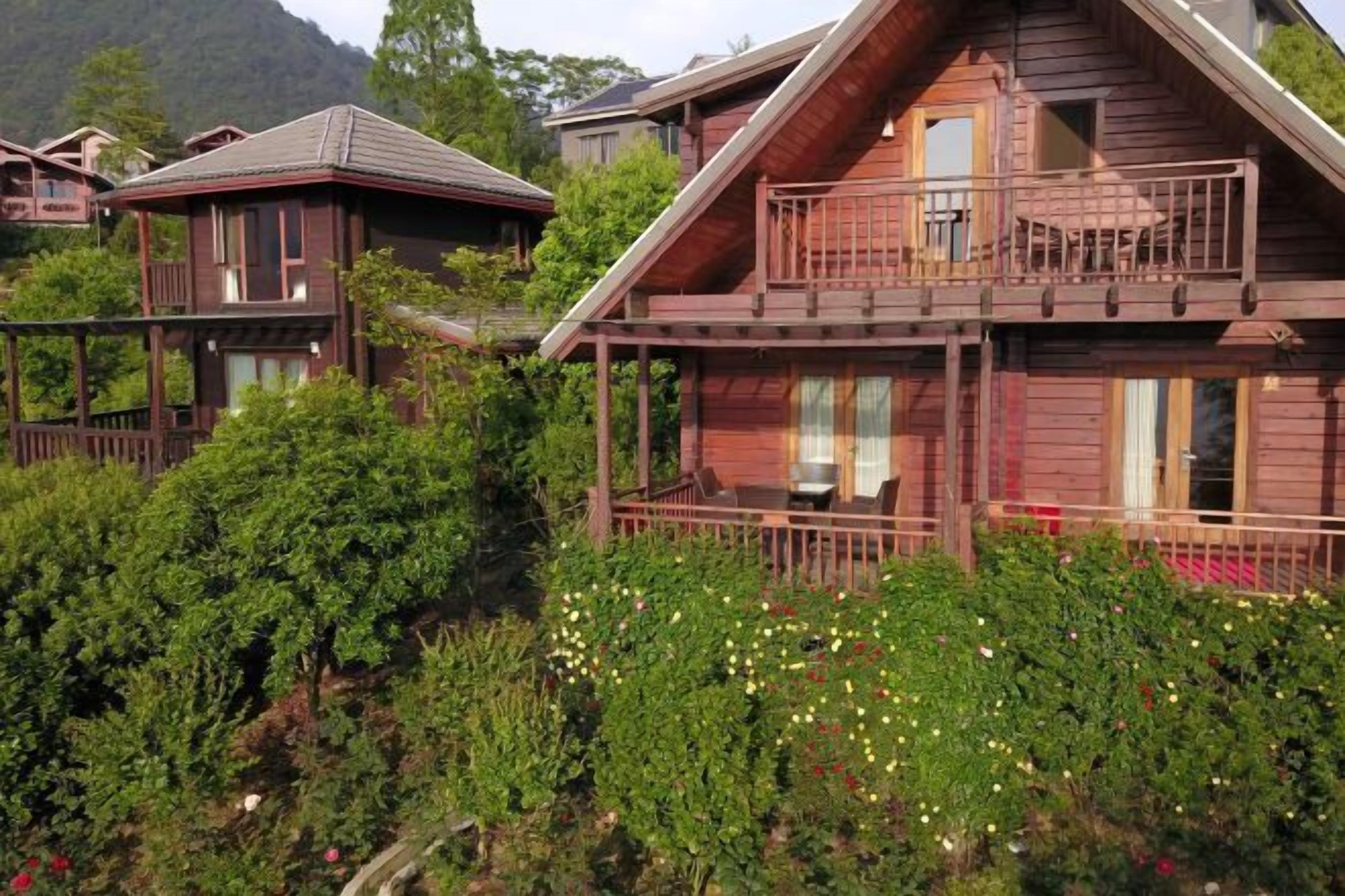 A wooden cabin with a balcony, surrounded by lush greenery and flowering plants.