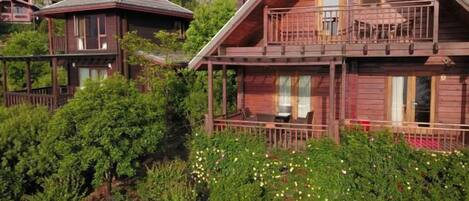 A wooden cabin with a balcony, surrounded by lush greenery and flowering plants.