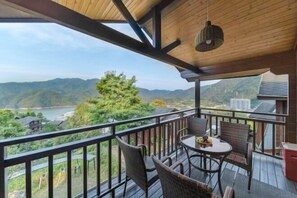 Un balcón con mobiliario de mimbre y una mesa, con vistas a un paisaje montañoso y una masa de agua.