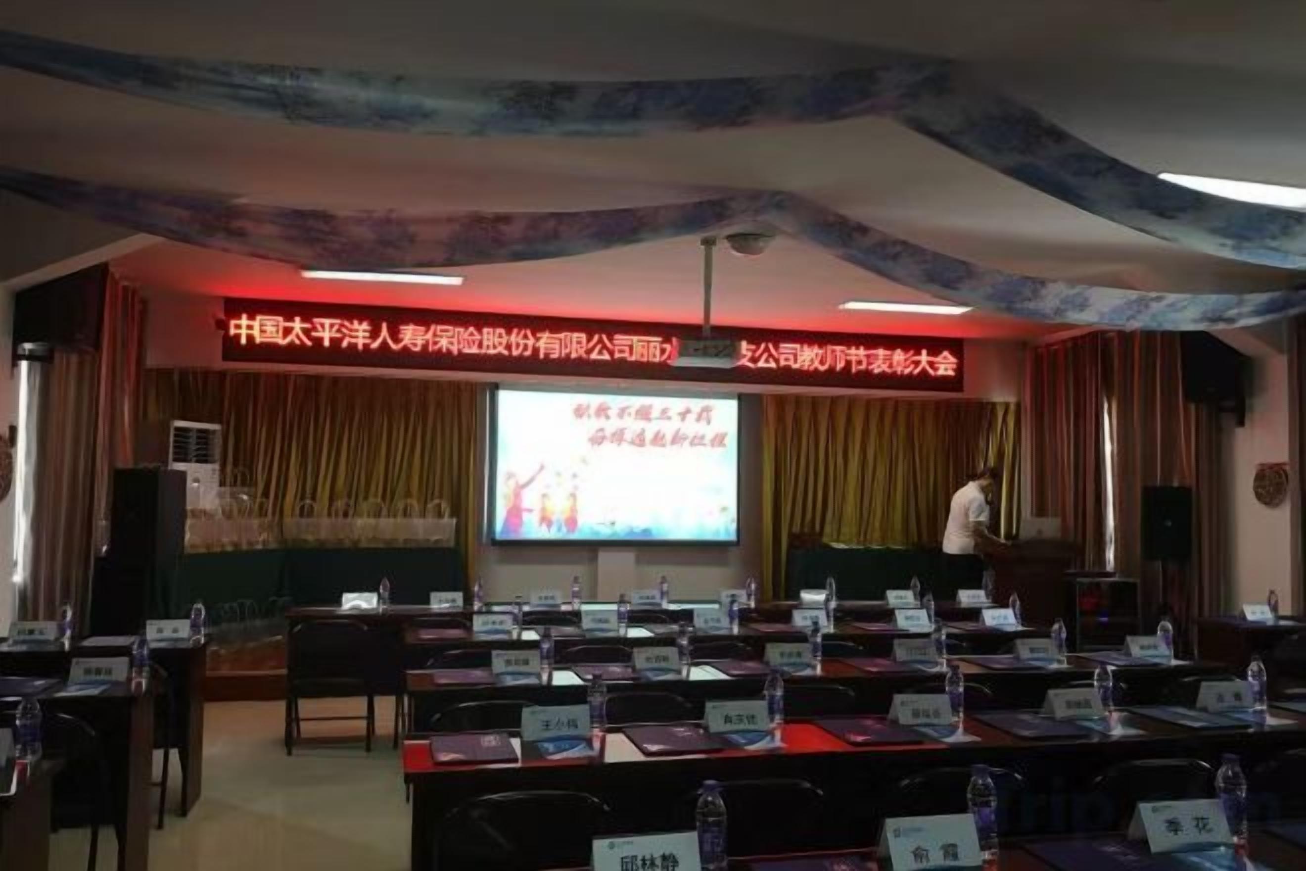 A conference room with a stage, a presentation screen, and rows of tables with nameplates.