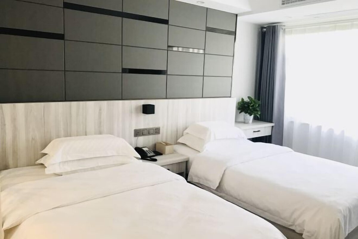 Una habitación de hotel con dos camas, una mesita de noche con un teléfono y una caja, un panel en la pared y una ventana con cortinas.
