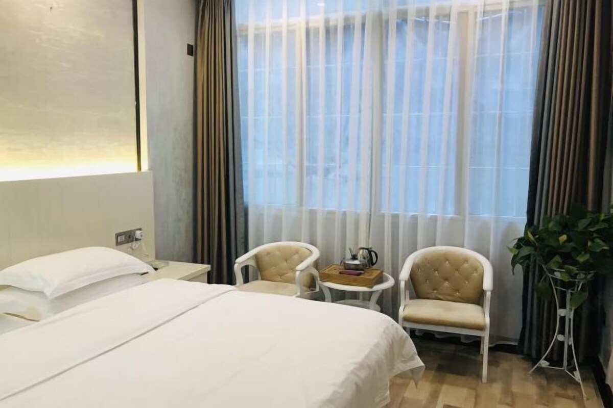 Habitación de hotel con una cama grande, dos sillones, una mesita con hervidor y una planta en maceta.