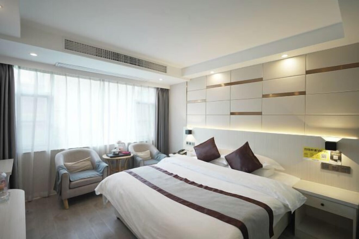 Una habitación de hotel moderna con una cama grande, una zona de estar con una silla y una mesita, y un ventanal con cortinas.
