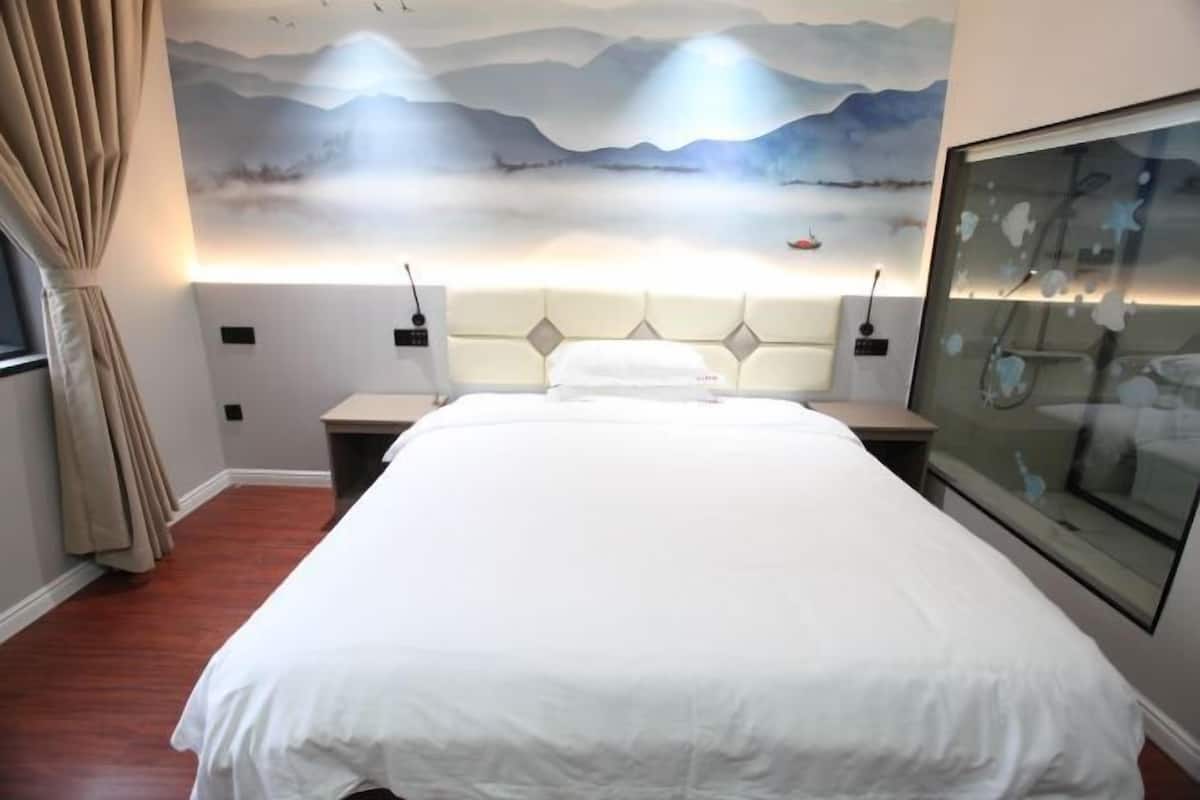 A hotel room with a large bed, a headboard with integrated lighting, a wall-mounted artwork, and a window with curtains.
