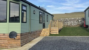 A green mobile home with a wooden deck and a stone wall in the background.