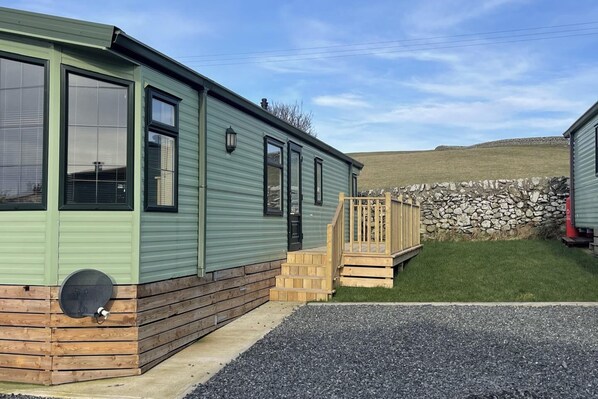 Una casa móvil verde con una terraza de madera y un muro de piedra al fondo.