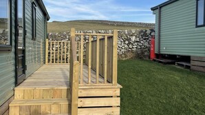 A wooden deck leading to a stone wall, with a grassy yard and a green building in the background.