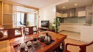 Une chambre d’hôtel moderne avec une table à manger en bois, préparée pour le thé, une télévision et un coin cuisine doté de meubles blancs.