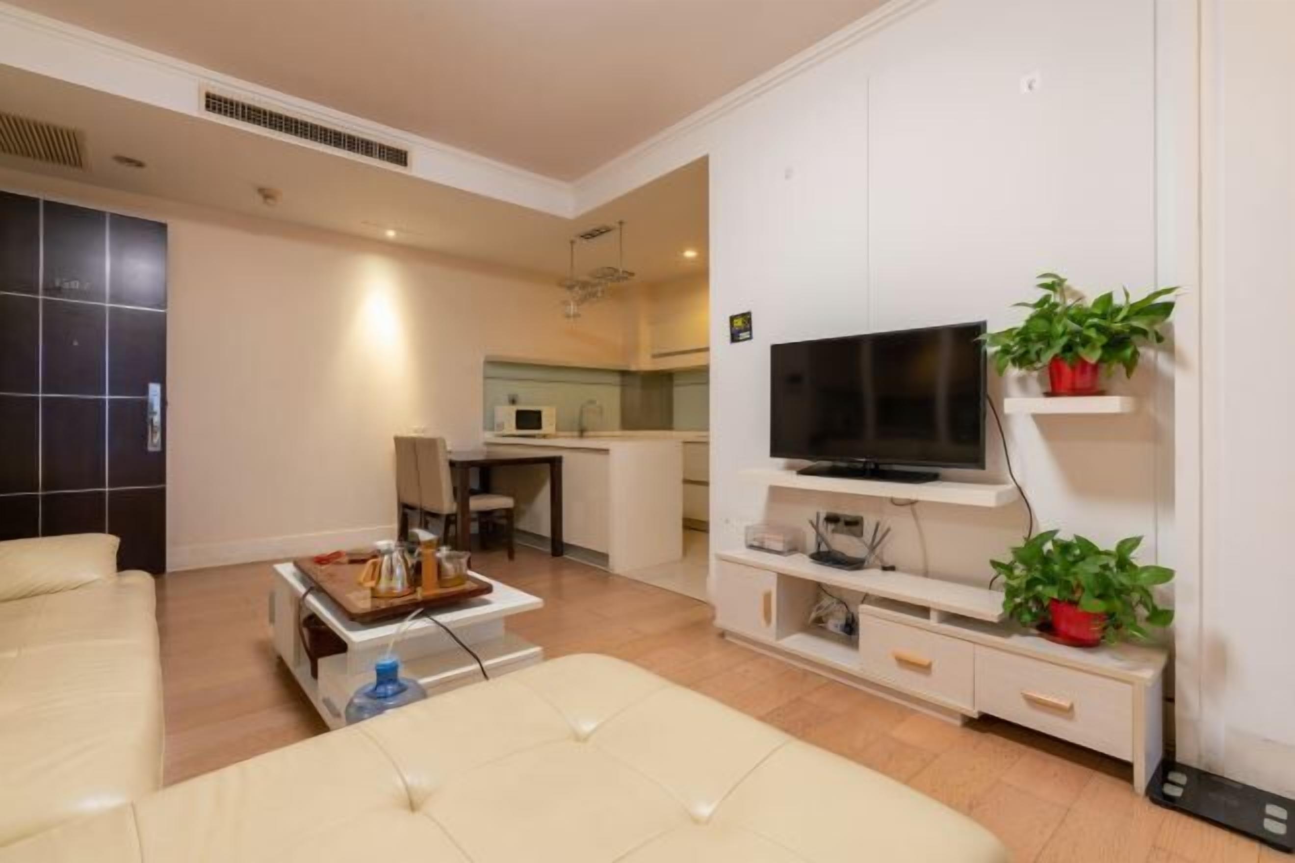 A modern living room with a sofa, a coffee table, and a TV on a white stand. There is a kitchen in the background with a microwave and a dining area.
