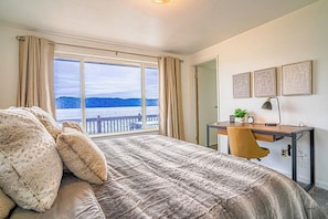 Een slaapkamer met een groot bed, een bureau met stoel, uitzicht op zee en drie foto's in lijsten aan de muur.