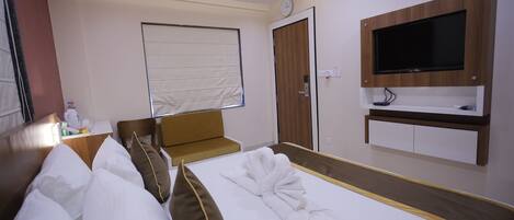 A hotel room with a neatly made bed, a television mounted on the wall, a desk with a chair, and a window with blinds.