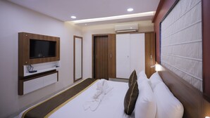A modern hotel room with a flat-screen TV mounted on the wall, a wooden wardrobe, a bed with white linens and a brown pillow, and an air conditioning unit above the wardrobe.