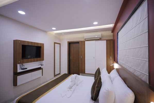 Una habitación de hotel moderna con un televisor de pantalla plana montado en la pared, un armario de madera, una cama con ropa blanca y una almohada marrón, y un equipo de aire acondicionado sobre el armario.