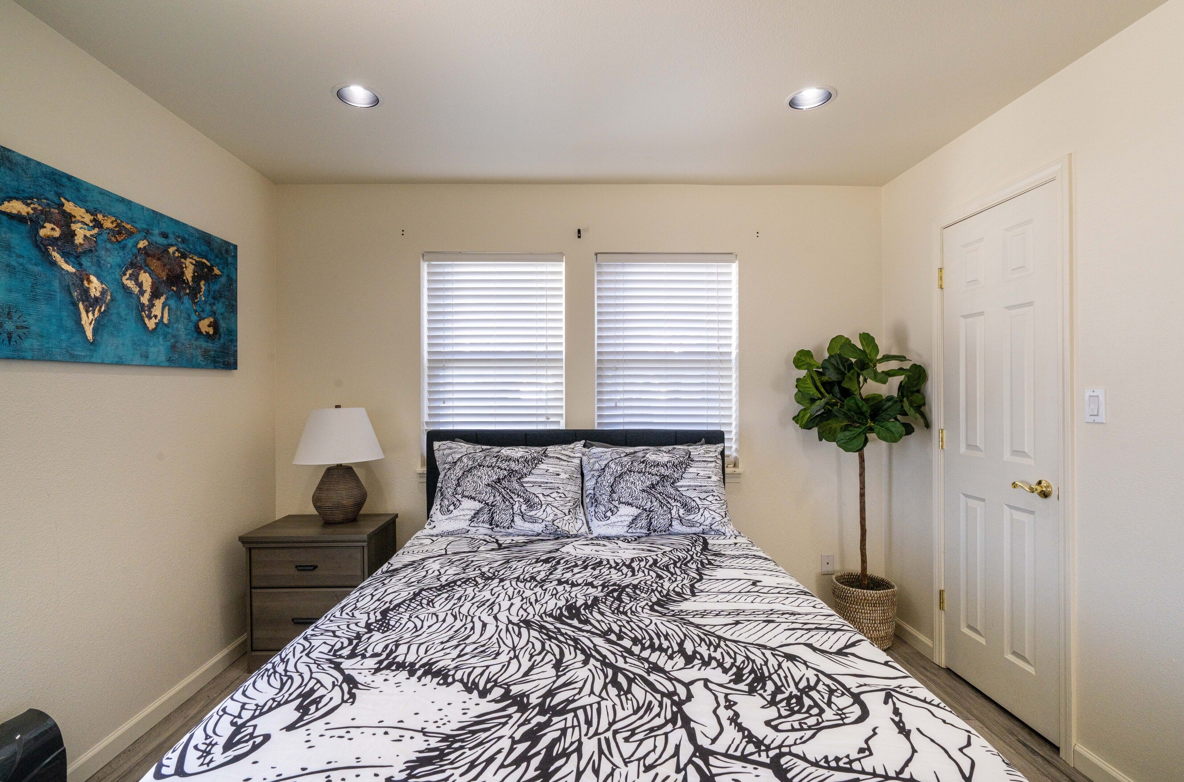 A bedroom with a bed, a world map on the wall, two windows with blinds, a nightstand with a lamp, and a potted plant.